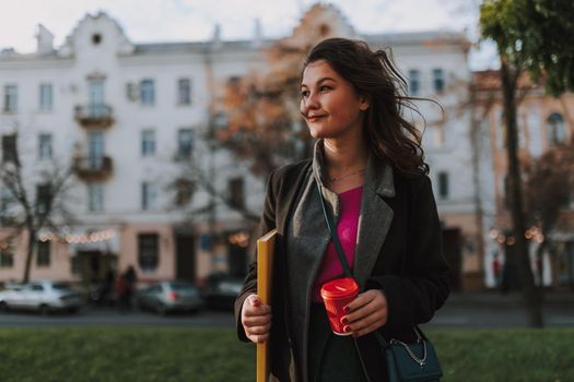 Waist up of happy beautiful woman walking in the city while holding hot drink and folder