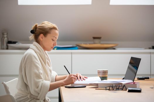 Young businesswoman wearing cosy warm bathrobe working remotly from home in winter Christmas time during 2020 corona virus pandemic. Work from home, selter in place, concept.