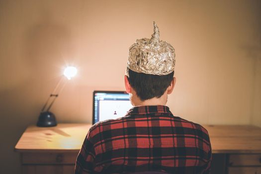 Young man with aluminum cap is sitting in the dark basement in front of a laptop. Conspiracy theory concept