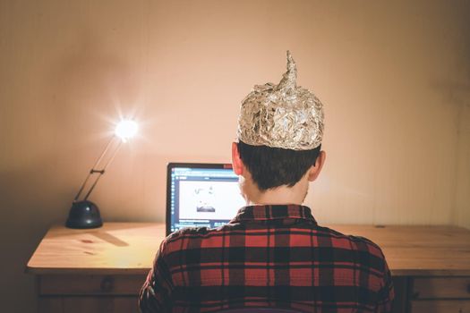 Young man with aluminum cap is sitting in the dark basement in front of a laptop. Conspiracy theory concept