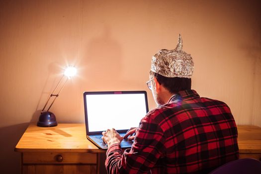 Young man with aluminum cap is sitting in the dark basement in front of a laptop. Conspiracy theory concept