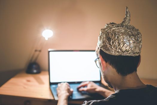 Young man with aluminum cap is sitting in the dark basement in front of a laptop. Conspiracy theory concept