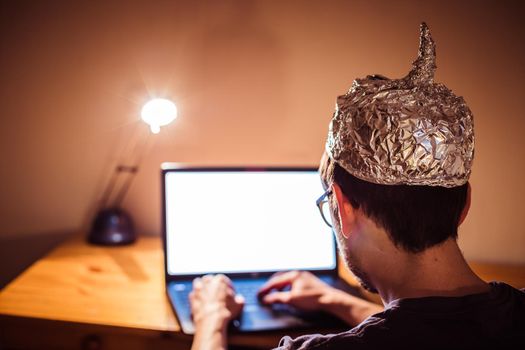 Young man with aluminum cap is sitting in the dark basement in front of a laptop. Conspiracy theory concept