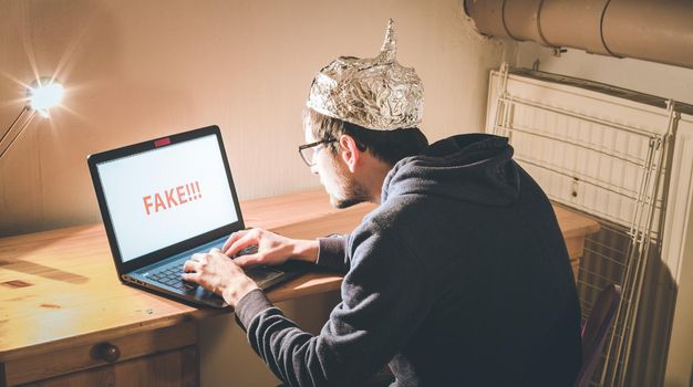 Young man with aluminum cap is sitting in the dark basement in front of a laptop. Conspiracy theory concept