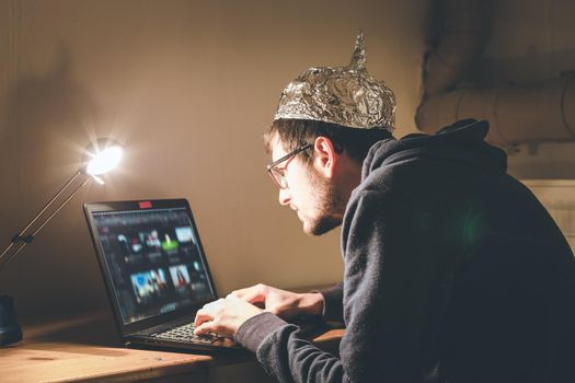 Young man with aluminum cap is sitting in the dark basement in front of a laptop. Conspiracy theory concept