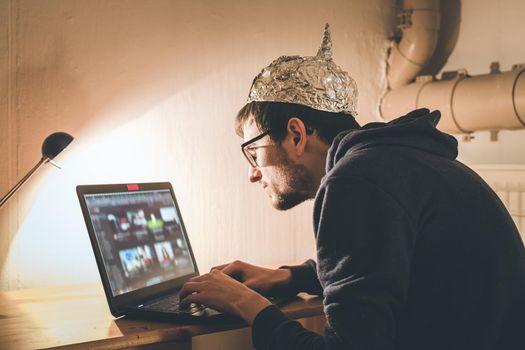 Young man with aluminum cap is sitting in the dark basement in front of a laptop. Conspiracy theory concept