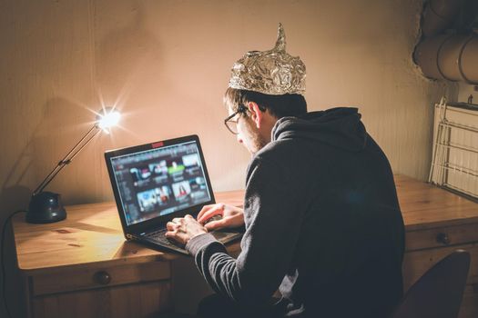 Young man with aluminum cap is sitting in the dark basement in front of a laptop. Conspiracy theory concept