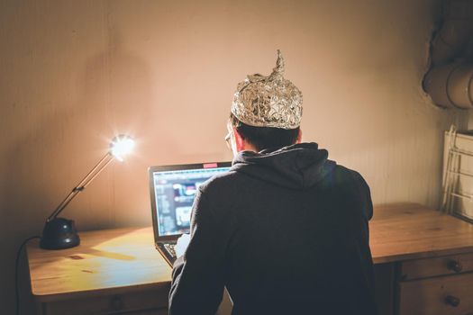 Young man with aluminum cap is sitting in the dark basement in front of a laptop. Conspiracy theory concept