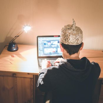 Young man with aluminum cap is sitting in the dark basement in front of a laptop. Conspiracy theory concept