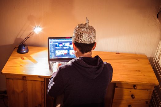 Young man with aluminum cap is sitting in the dark basement in front of a laptop. Conspiracy theory concept