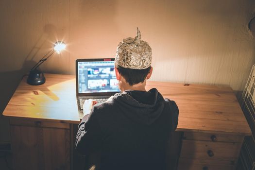 Young man with aluminum cap is sitting in the dark basement in front of a laptop. Conspiracy theory concept