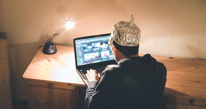 Young man with aluminum cap is sitting in the dark basement in front of a laptop. Conspiracy theory concept