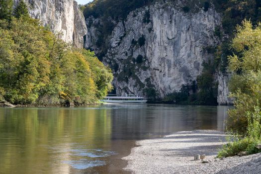 Passenger ship passing Danube breakthrough near Kelheim, Bavaria, Germany in autumn with gravel bank in foreground and limestone formations in the background