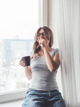 Woman with eyeglasses and curly hair sits on windowsill. Sleepy woman yawns. Morning cup of hot coffee.