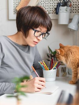 Woman with short hair cut is drawing in notebook. Cute ginger cat sits near her. Fluffy pet and artist. Calming hobby, anti stress leisure.