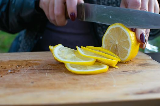 women's hands cut a juicy lemon with a knife on a wooden board in a summer cottage in the open air