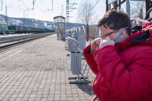 a man waiting for a train on the platform of a railway station adjusts a medical anti-bacterial anti-virus mask on his face