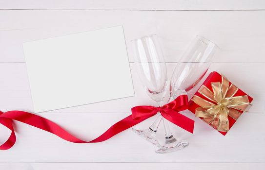 Valentine day concept, wineglass and red ribbon and gift box with greeting card on white wooden table background, champaign glass and postcard, top view, couples wine glass together, holiday concept.