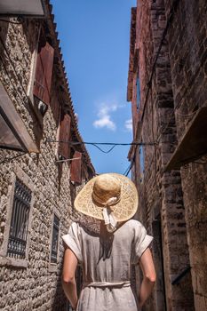 Rear view of beautiful blonde young female traveler wearing straw sun hat sightseeing and enjoying summer vacation in an old traditional costal town at Adriatic cost, Croatia.