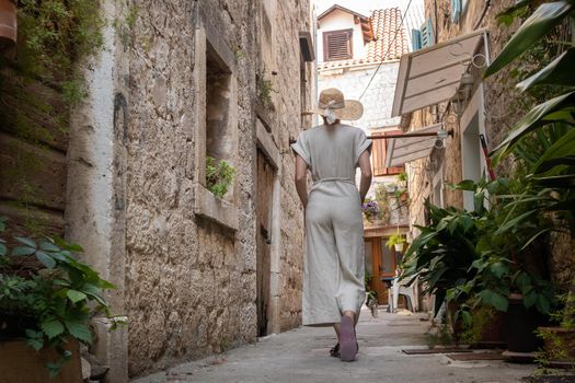 Rear view of beautiful blonde young female traveler wearing straw sun hat sightseeing and enjoying summer vacation in an old traditional costal town at Adriatic cost, Croatia.