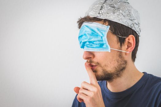 Young angry man with face mask over the eyes and aluminum hat is doing a psst! gesture