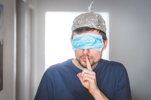 Young angry man with face mask over the eyes and aluminum hat is doing a psst! gesture