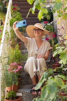 Beautiful female tourist wearing big straw sun hat and sundress sitting and relaxing on old stone house stairs during summer travel on Mediterranean cost on hot summer day and taking selfie..