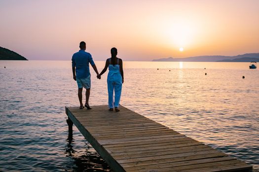 a couple seated on a wooden jetty, looking at colorful sunset on the sea, men, and women watching a sunset in Crete Greece. Europe