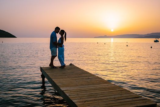 a couple seated on a wooden jetty, looking at colorful sunset on the sea, men, and women watching a sunset in Crete Greece. Europe