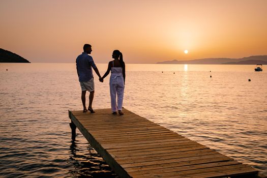 a couple seated on a wooden jetty, looking at colorful sunset on the sea, men, and women watching a sunset in Crete Greece. Europe