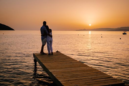 a couple seated on a wooden jetty, looking at colorful sunset on the sea, men, and women watching a sunset in Crete Greece. Europe