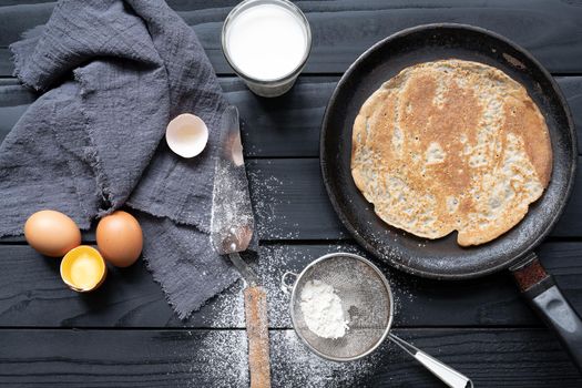 Hot pancake in black pan on black table with flour, milk and eggs.