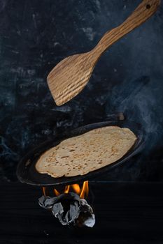 Hot pancake in black pan on black table with flour, milk and eggs.