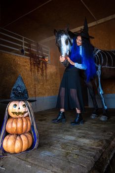 A girl in a witch costume hugs a horse in a corral, in the foreground an evil figure of pumpkins