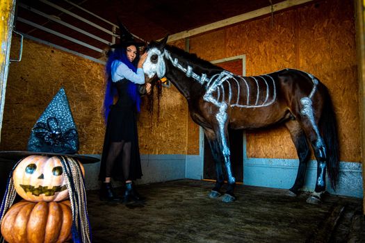 A girl in a witch costume holds a horse by the bridle in a corral on a farm, a skeleton is painted on a horse in white paint