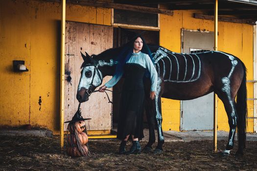 A girl dressed as a witch on a farm, a horse with a skeleton is drawn next to it, as well as three pumpkins in the shape of an evil man