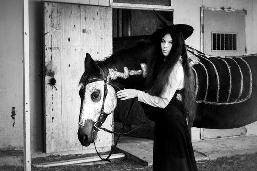 Black and white portrait of a girl in a witch costume with a horse on which a skeleton is drawn