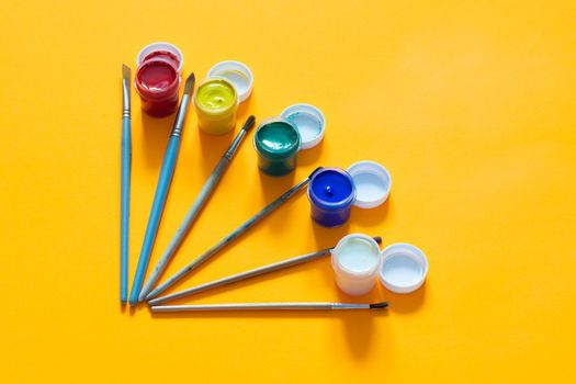 Brushes and jars of gouache are laid out on a yellow background