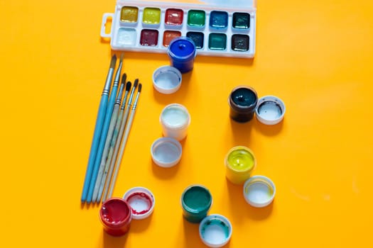 Brushes, jars of gouache and watercolor paints lie on a yellow background