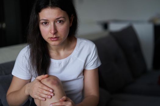 Portrait of beaten woman with tears of her face and bruise on knee sit on the sofa.