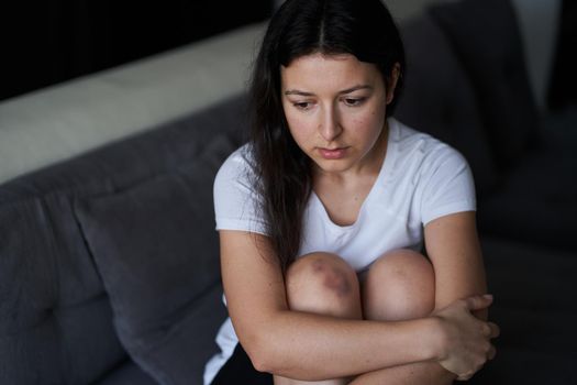 Portrait of beaten woman with tears of her face and bruise on knee sit on the sofa.