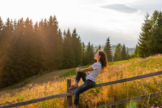 The girl enjoy sunset sitting on wooden fence on highlands field next to forest.