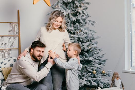 Christmas Family Happiness Portrait of dad, pregnant mom and little son sitting armchair at home near Christmas tree hug smile European young adult family holiday morning