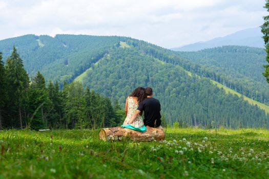 Young couple in love hugs sitting on the log and enjoy betiful of nature mountain lanscape overgrov with green trees.