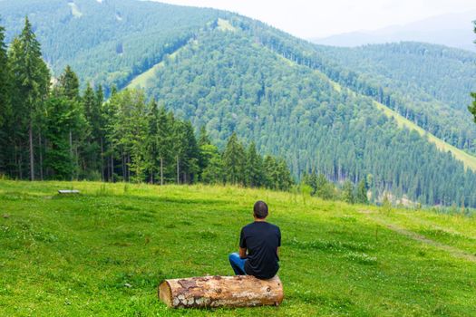 Guy sitting on log and enjoy peaceful green mountains landscape. Peace of mind and relax.