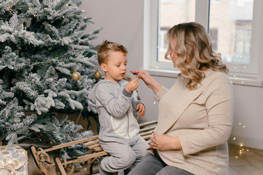 Holiday Christmas Caucasian mother playing with little cute son near decorated New year tree at home Family tradition