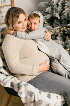 Mom plays with child. Happy family Portrait In Home pregnant mother embraces his little son. Happy new year. decorated Christmas tree Christmas morning bright living room Caucasian woman with child