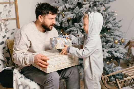 Holiday Christmas Handsome father playing with small cute son near decorated New year tree at home Family tradition boy gives a gift to his father