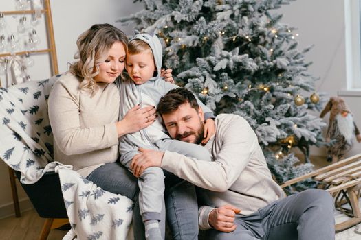 Christmas Family Happiness Portrait of dad, pregnant mom and little son sitting armchair at home near Christmas tree hug smile European young adult family holiday morning