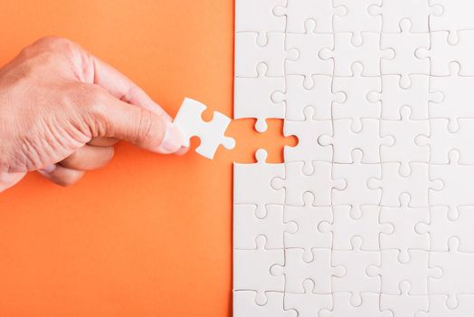 Top view flat lay of hand-holding last piece white paper jigsaw puzzle game last pieces put to place for solve problem complete mission, studio shot on an orange background, quiz calculation concept